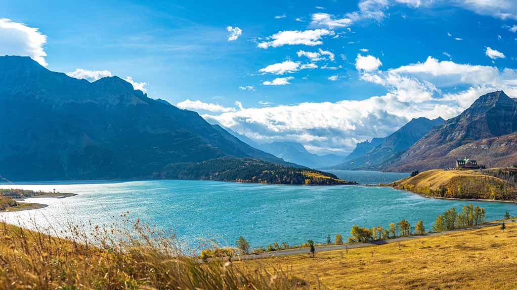 Wide landscape view of Waterton Lake with blue water, rolling shoreline, and rugged mountains under a bright sky in Waterton Lakes National Park